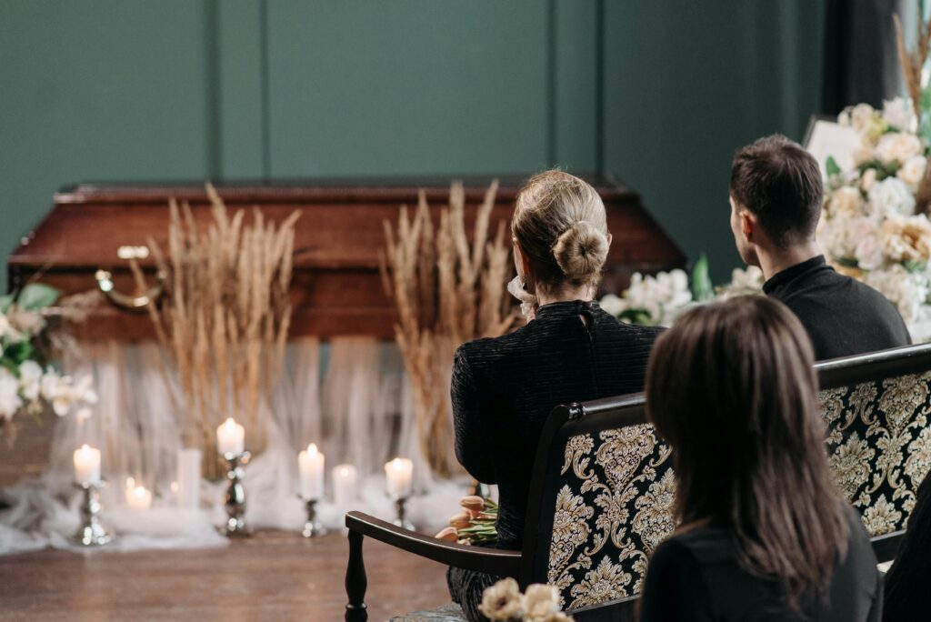 A respectful gathering of family and friends at a funeral service indoors, with floral arrangements and candles.